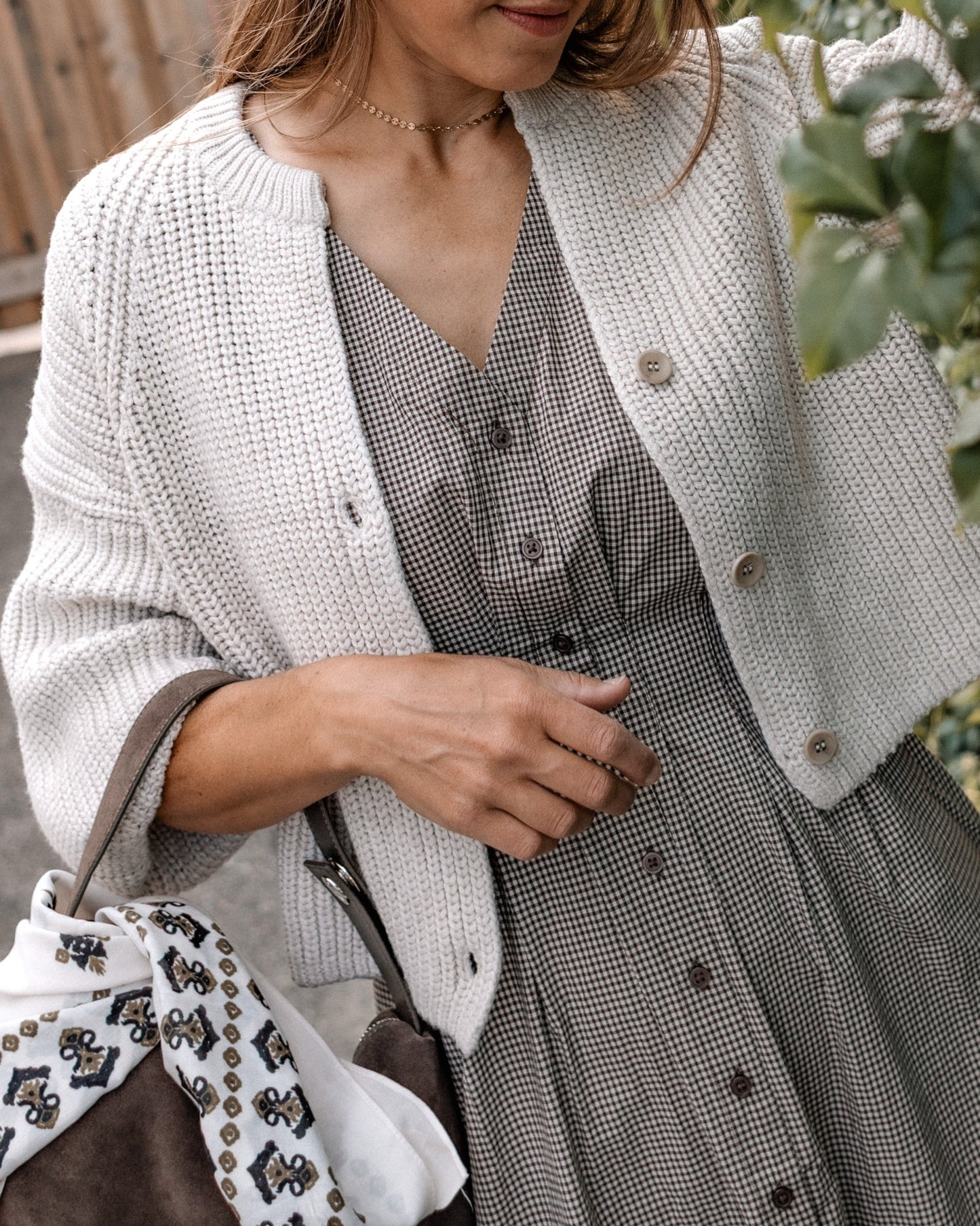 A woman wearing a light gray knit cardigan over a gray patterned button-front dress, carrying a brown handbag with a white patterned scarf, outdoors near greenery.